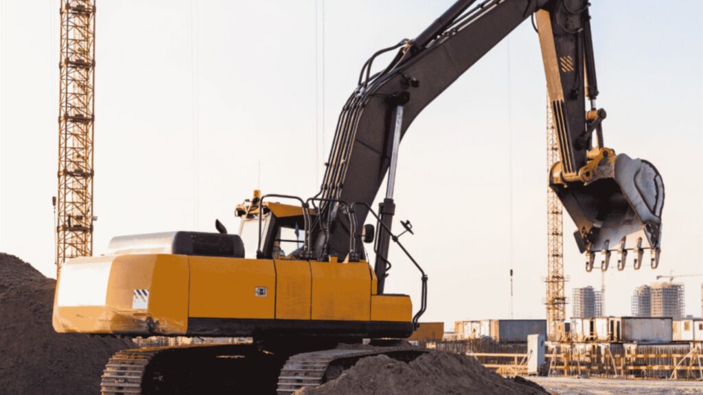 A large orange tracked excavator with its arm and bucket raised on a construction site, with tower cranes and buildings under construction behind.