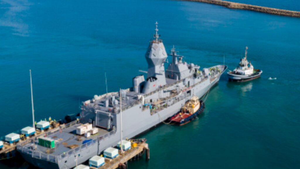 A grey naval warship berthed alongside a port wharf with a tug boat assisting under a blue sky.