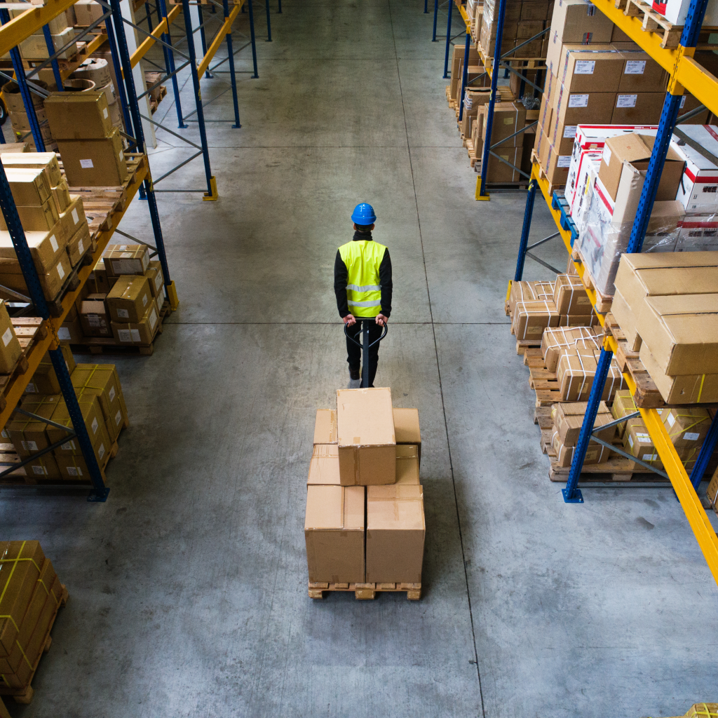 A warehouse worker in a hi-vis vest pulling a loaded pallet jack down an aisle between storage racks.