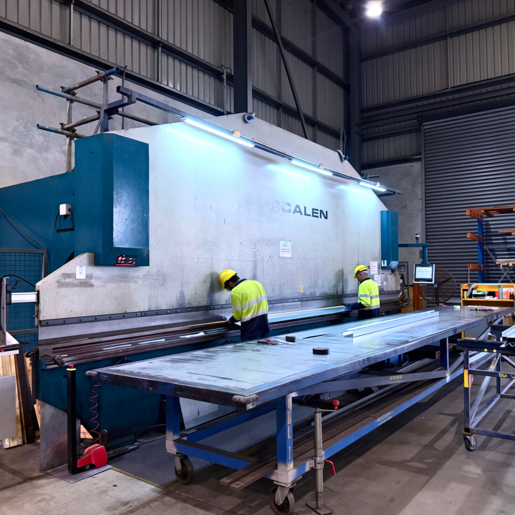 Two workers in hi-vis and hard hats operating a large press brake to bend sheet metal.