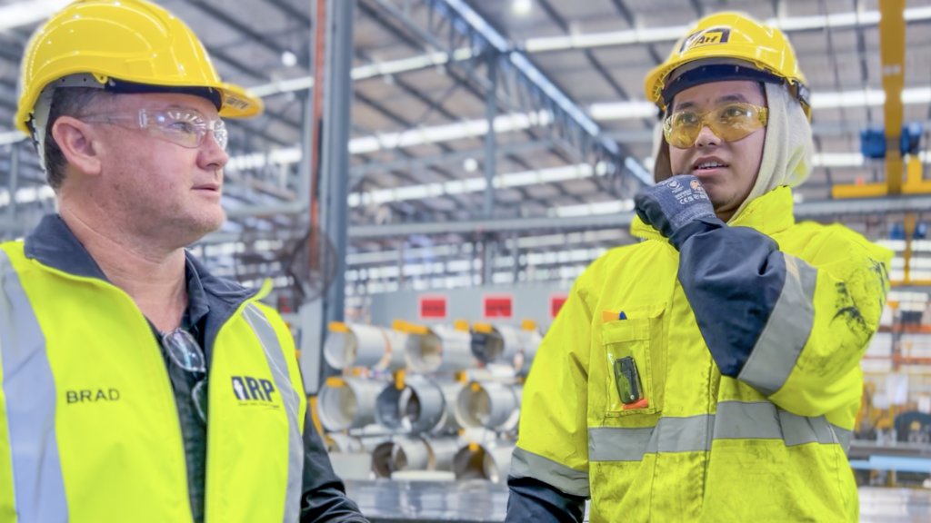 Two workers in hi-vis and hard hats talking on a BlueScope factory floor beside steel coils.