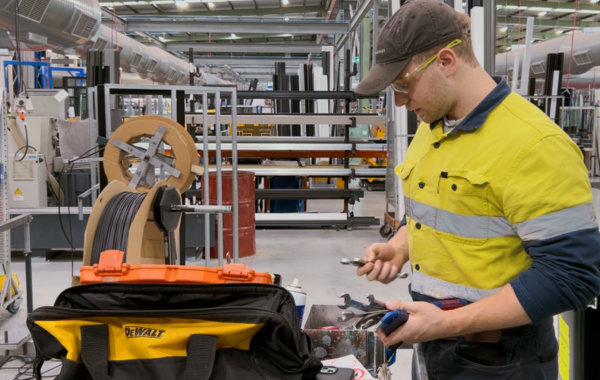 A male warehouse worker with blonde hair wearing a black cap, yellow safety glasses and yellow hi-vis holding a variety of tools such as spanners in his hand. 
