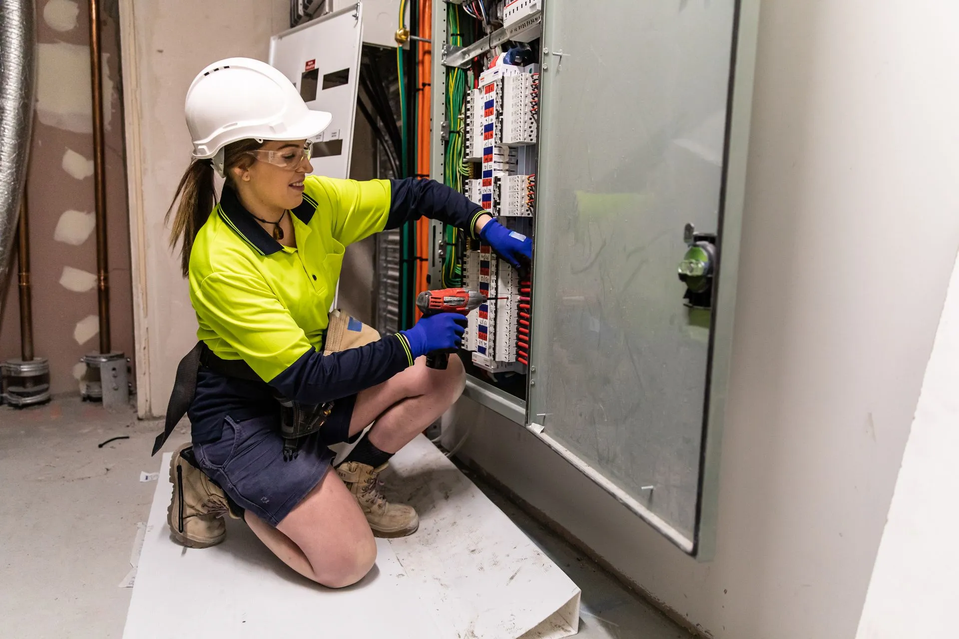 A female electrician in hi-vis and a hard hat wiring an electrical distribution board.